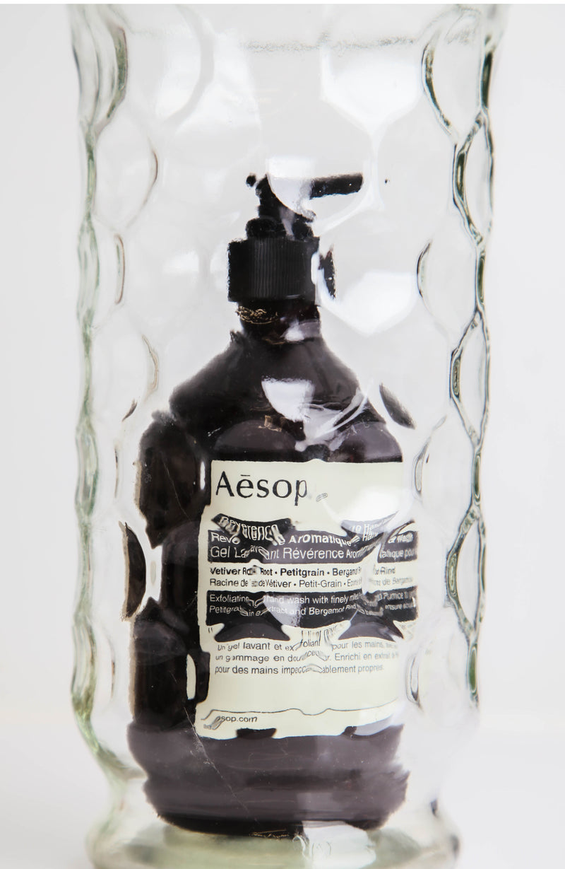 Aesop bottle inside a clear sculptural vase with white background 