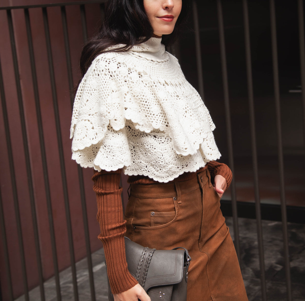 model wearing crochet cape with brown faux leather skirt, brown long sleeve knit top and grey bag