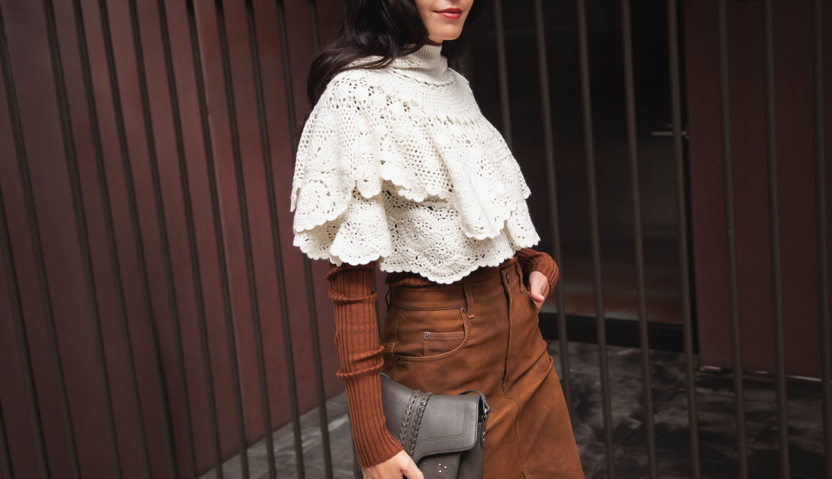 model wearing crochet cape with brown faux leather skirt, brown long sleeve knit top and grey bag