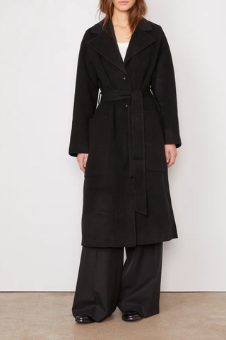 front profile model wearing Carlita Coat Black wool Single breasted long coat. Three covered buttons. Slightly oversized. Tailored collar. Raglan sleeves. Patch pockets on front. Single vent at center back. Self fabric belt.