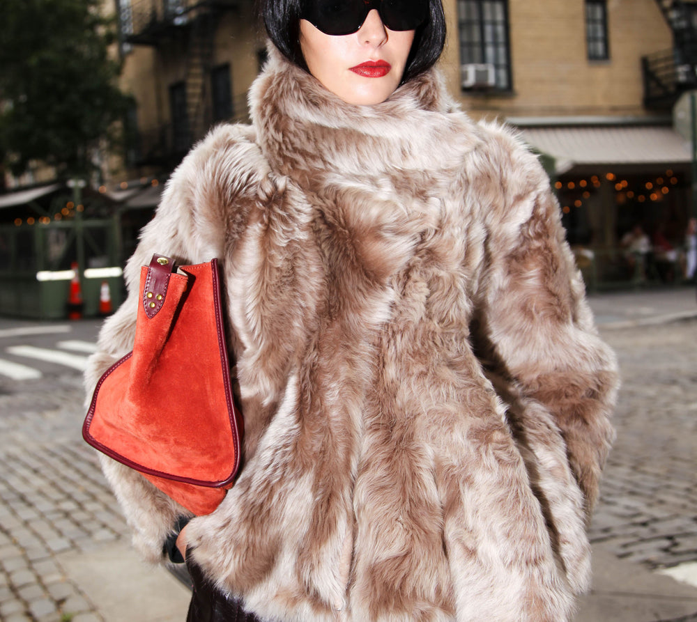 model wearing black sunglasses, beige fur coat and red suede bag, with a corner restaurant yellow background