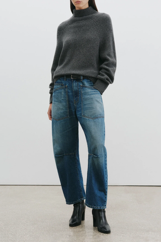front profile mode wearing Shon Jeans Bedford mid blue Wash Mid-rise, relaxed fit barrel leg jean. Uniquely curved silhouette. Seam detail at knees.