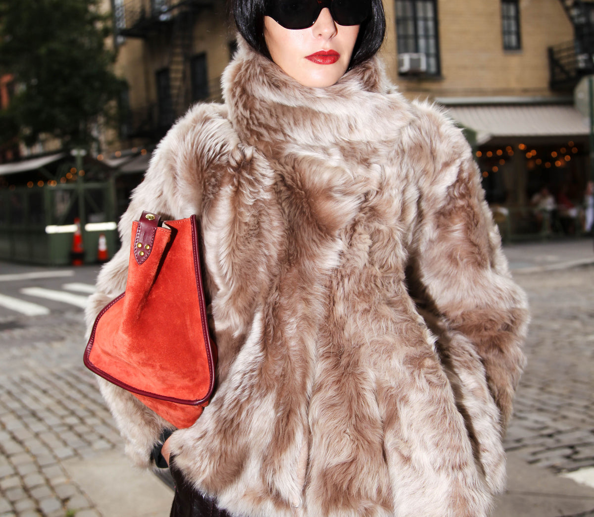 model wearing black sunglasses, beige fur coat and red suede bag with a street corner and restaurant background